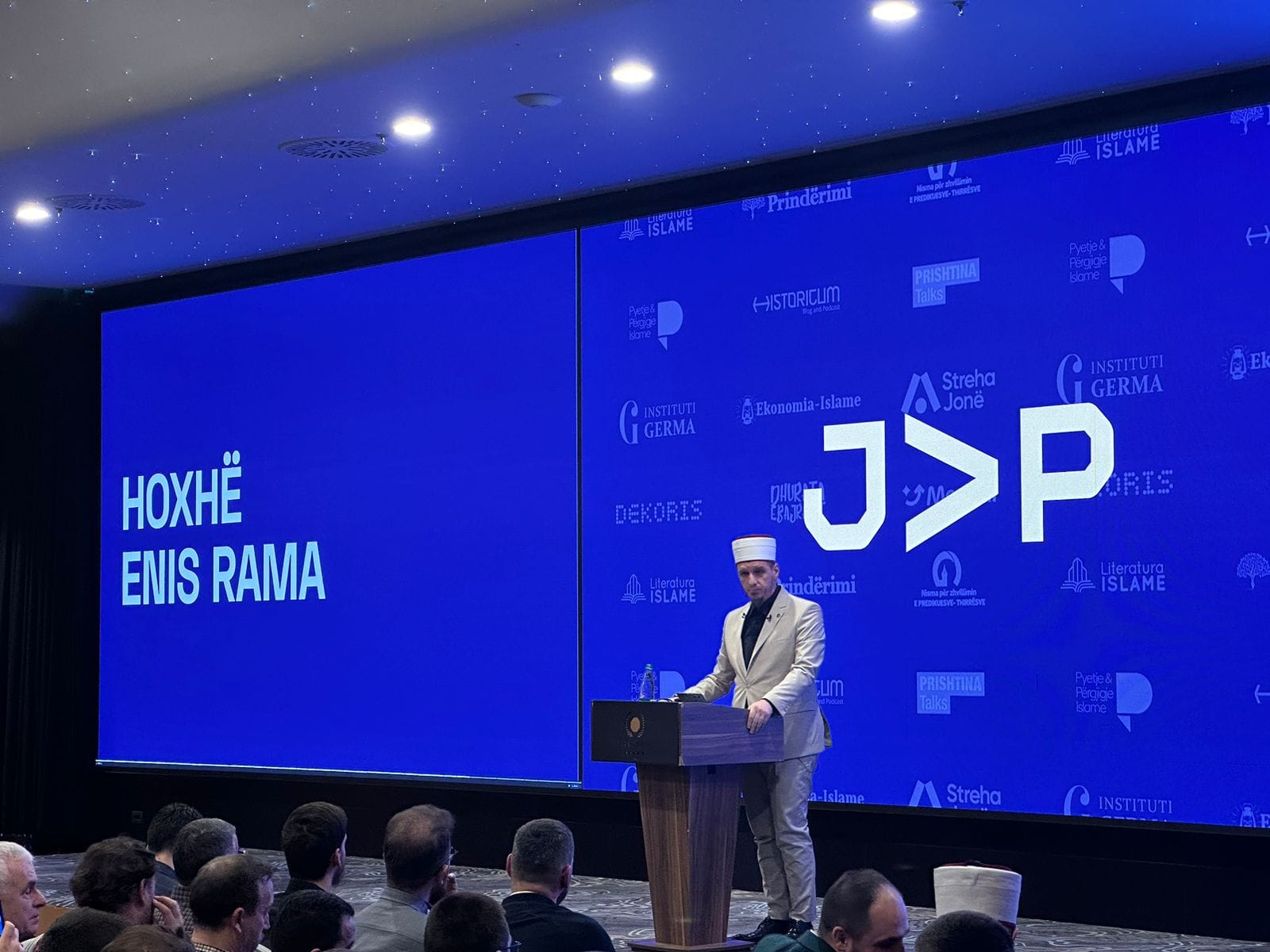 Folësi në podium para ekranit të madh me emrin e tij, në sallën e mbushur