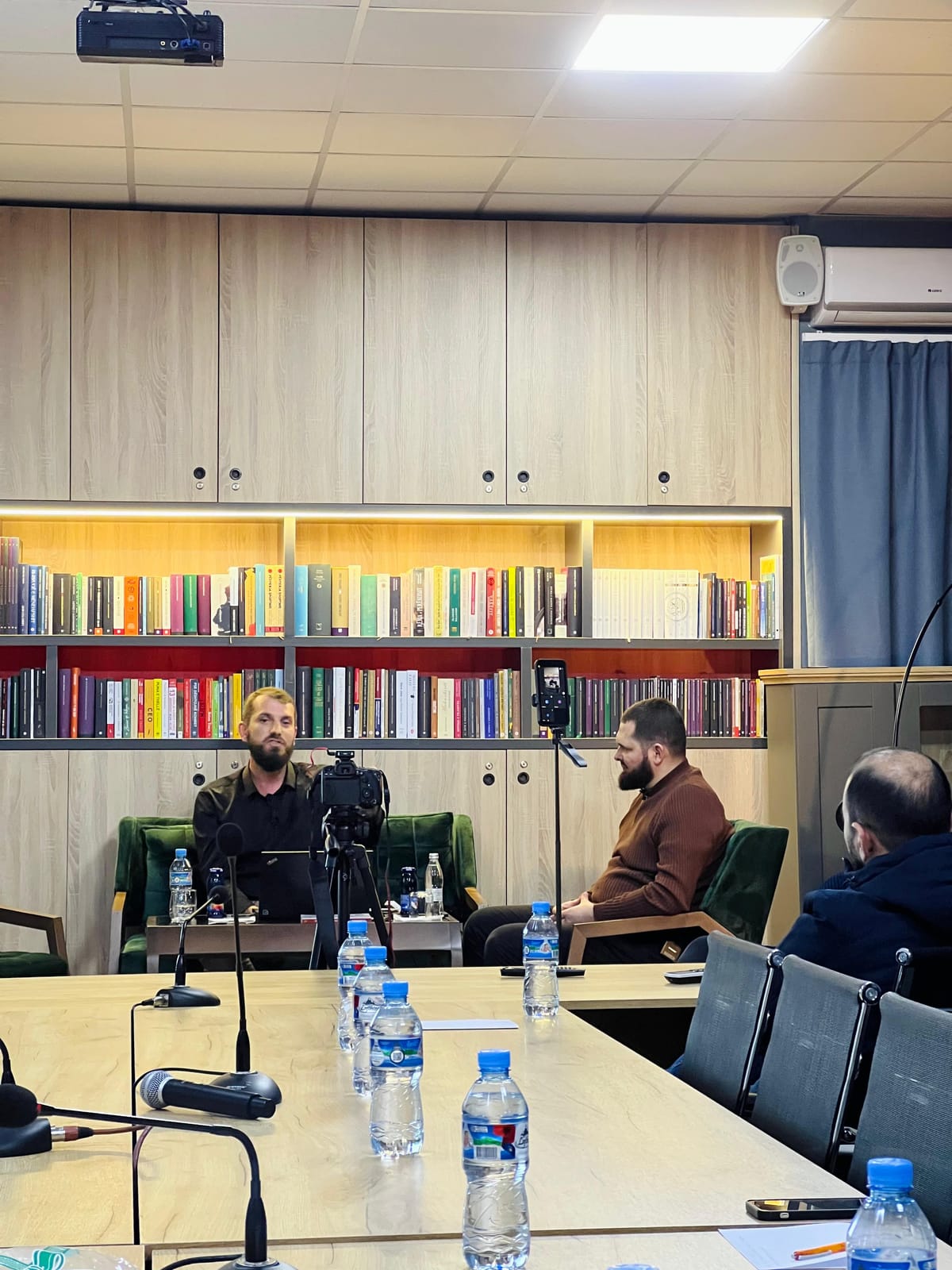 Dy folës me mikrofona rreth tryezës, me rafte librash në sfond