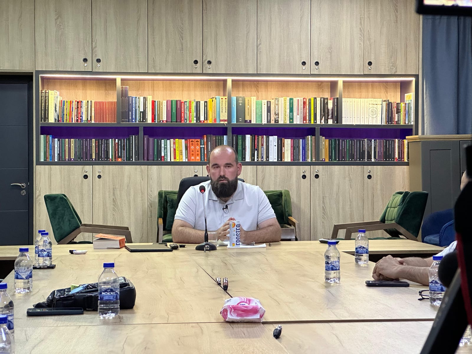 Folësi i ulur pas tryezës me mikrofon, me rafte librash në sfond