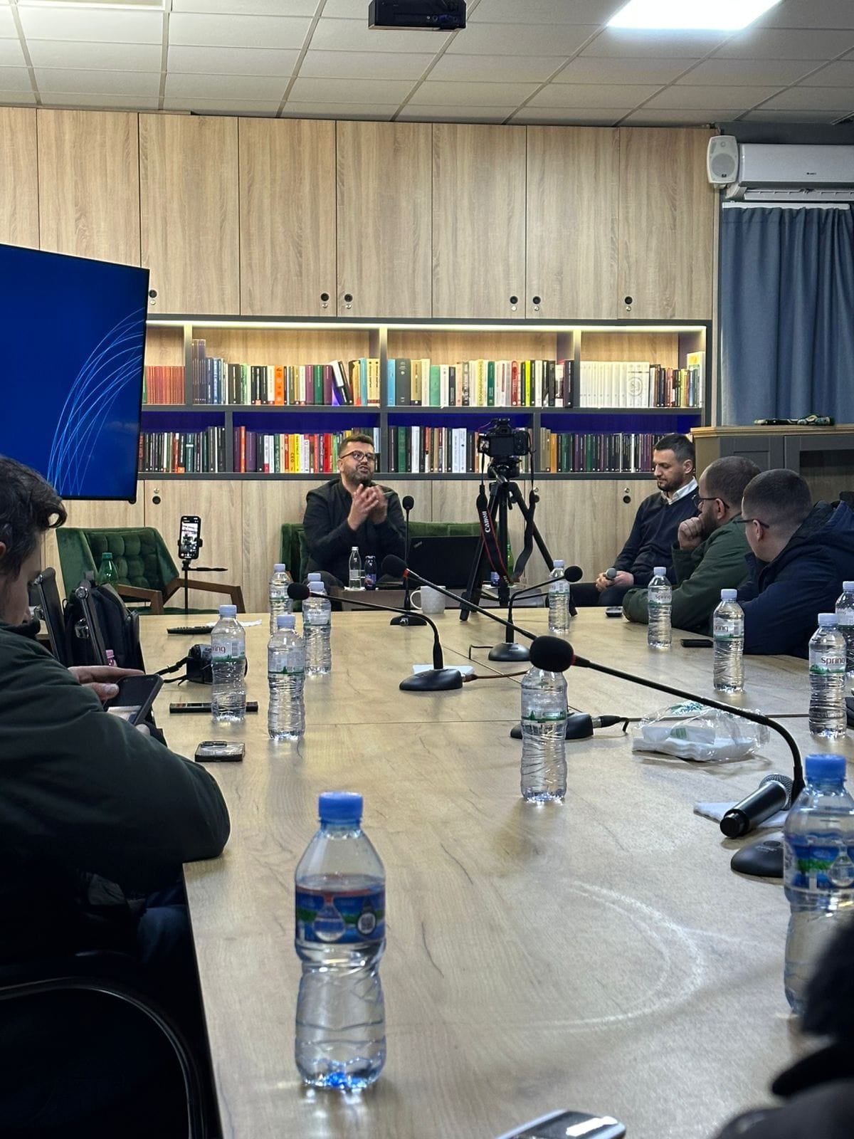 Folësi në bisedë me pjesëmarrësit rreth tryezës, me kamerë përpara