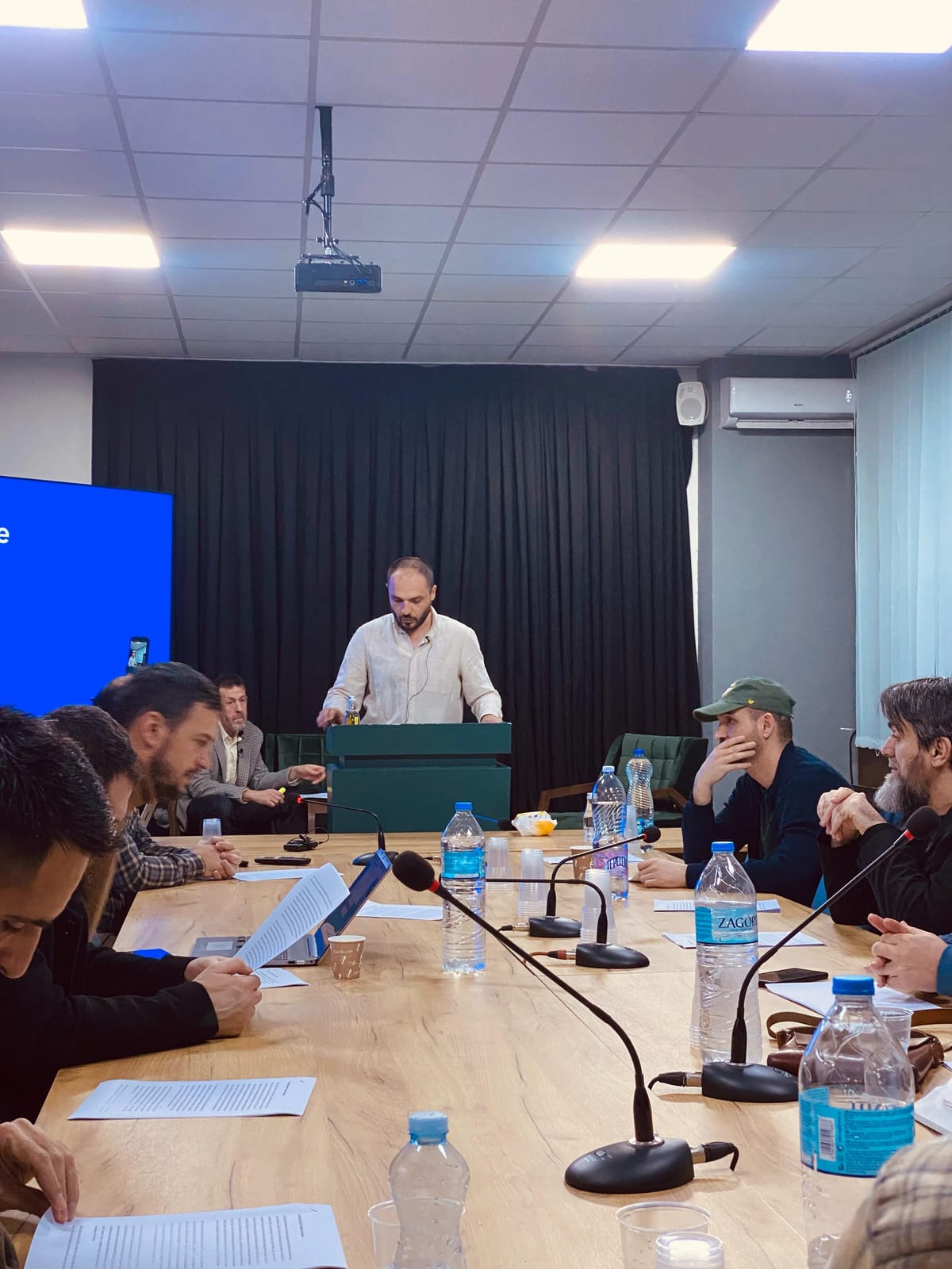 Folësi në këmbë pas podiumit me mikrofon, me ekran blu në sfond
