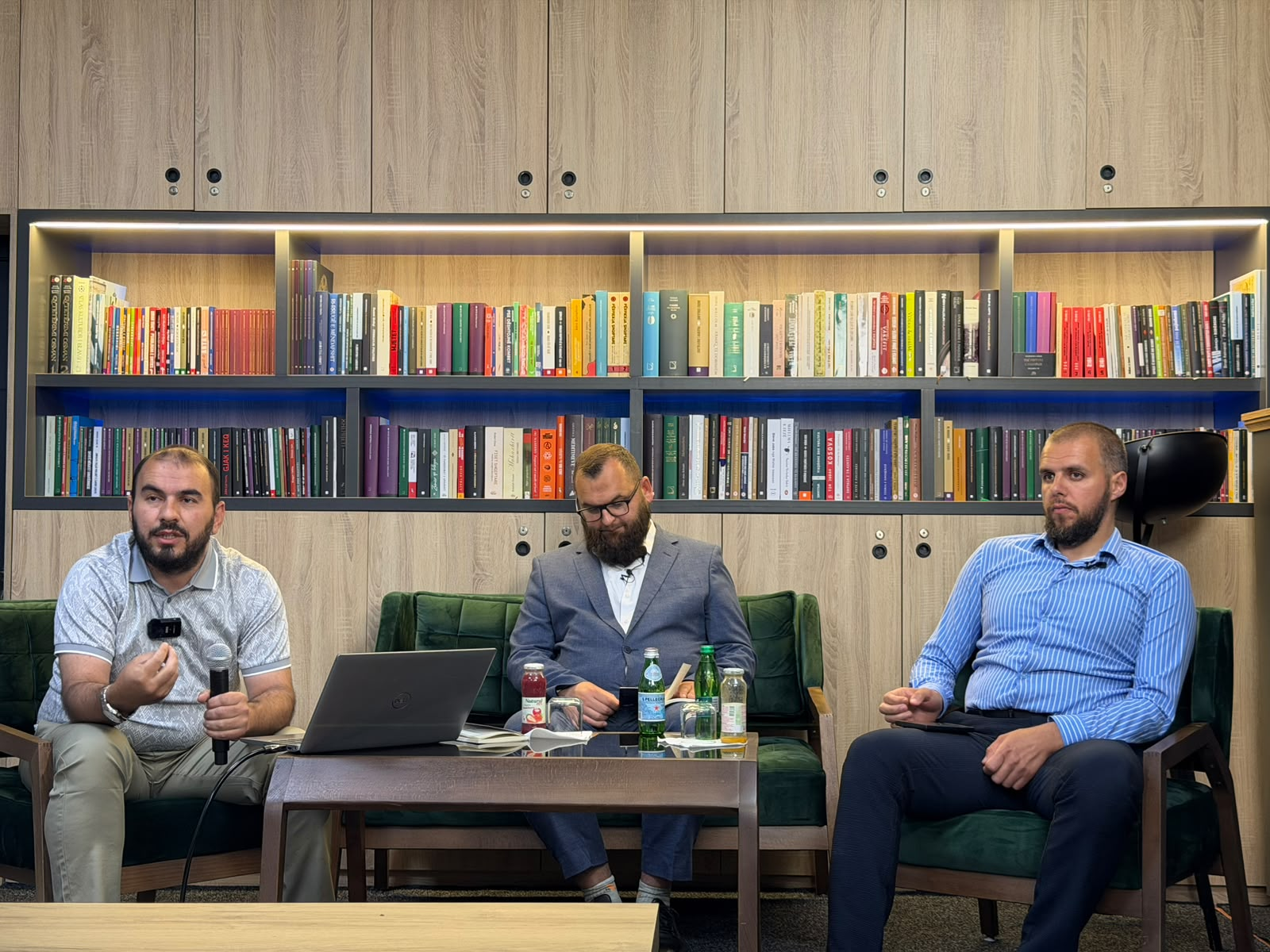 Tre folës të ulur në panel me mikrofona, para rafteve me libra gjatë një diskutimi